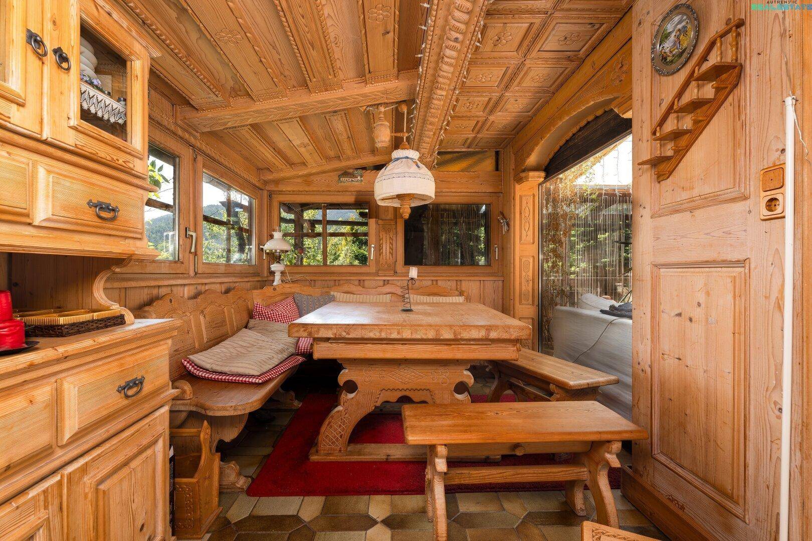 Rustikaler Essbereich im Wintergarten mit Holztisch, Bänken und geschnitzten Details.