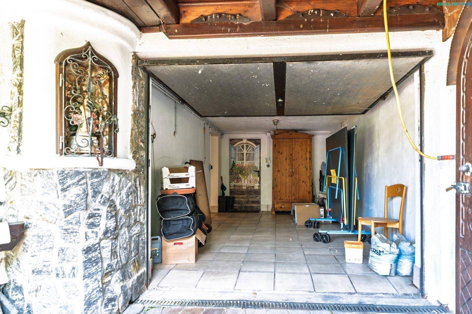 Offene Garage mit Fliesenboden, Steinwand und Stauraum für verschiedene Gegenstände.