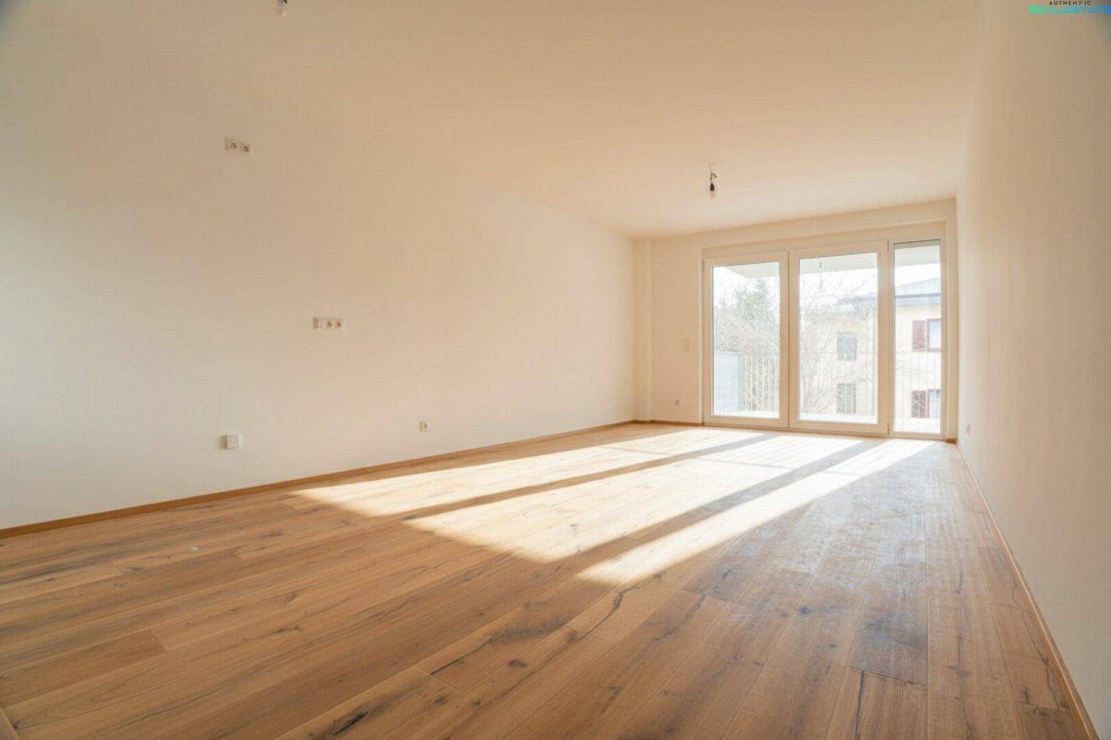 Leeres, lichtdurchflutetes Zimmer mit Holzboden und großen Fenstern zum Balkon.