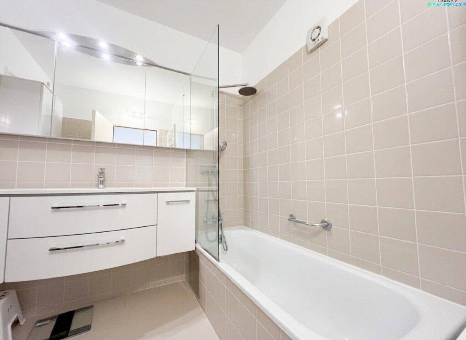 Helles Badezimmer mit beigefarbenen Fliesen, Badewanne mit Duschfunktion und Waschtischunterschrank.