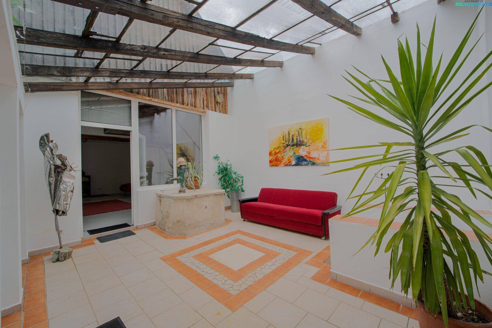 Überdachte Terrasse mit Fliesenboden, Sitzgelegenheiten und einer großen Yucca-Pflanze.