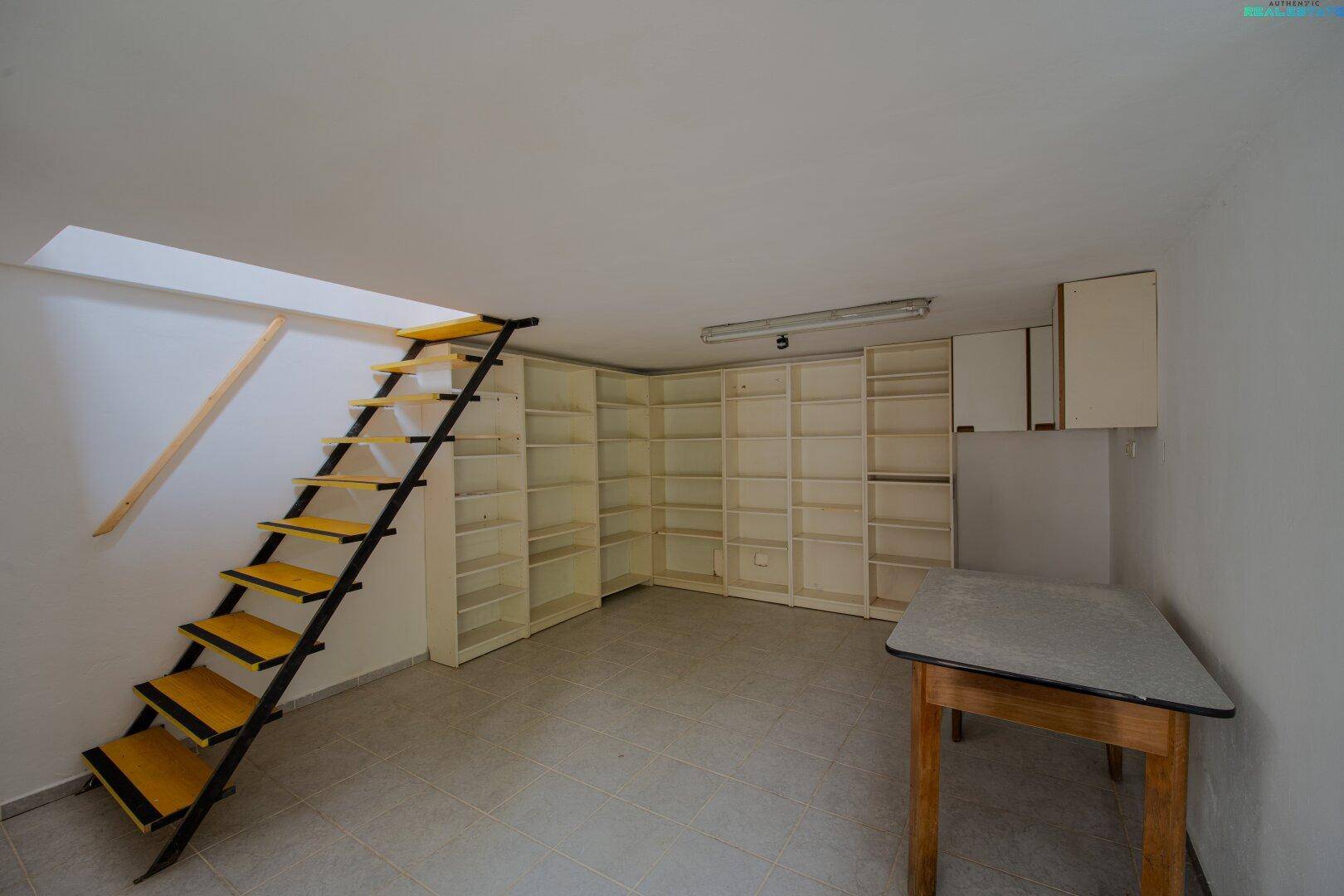 Lagerraum mit Regalen, Arbeitstisch und einer Treppe zu einem oberen Bereich.