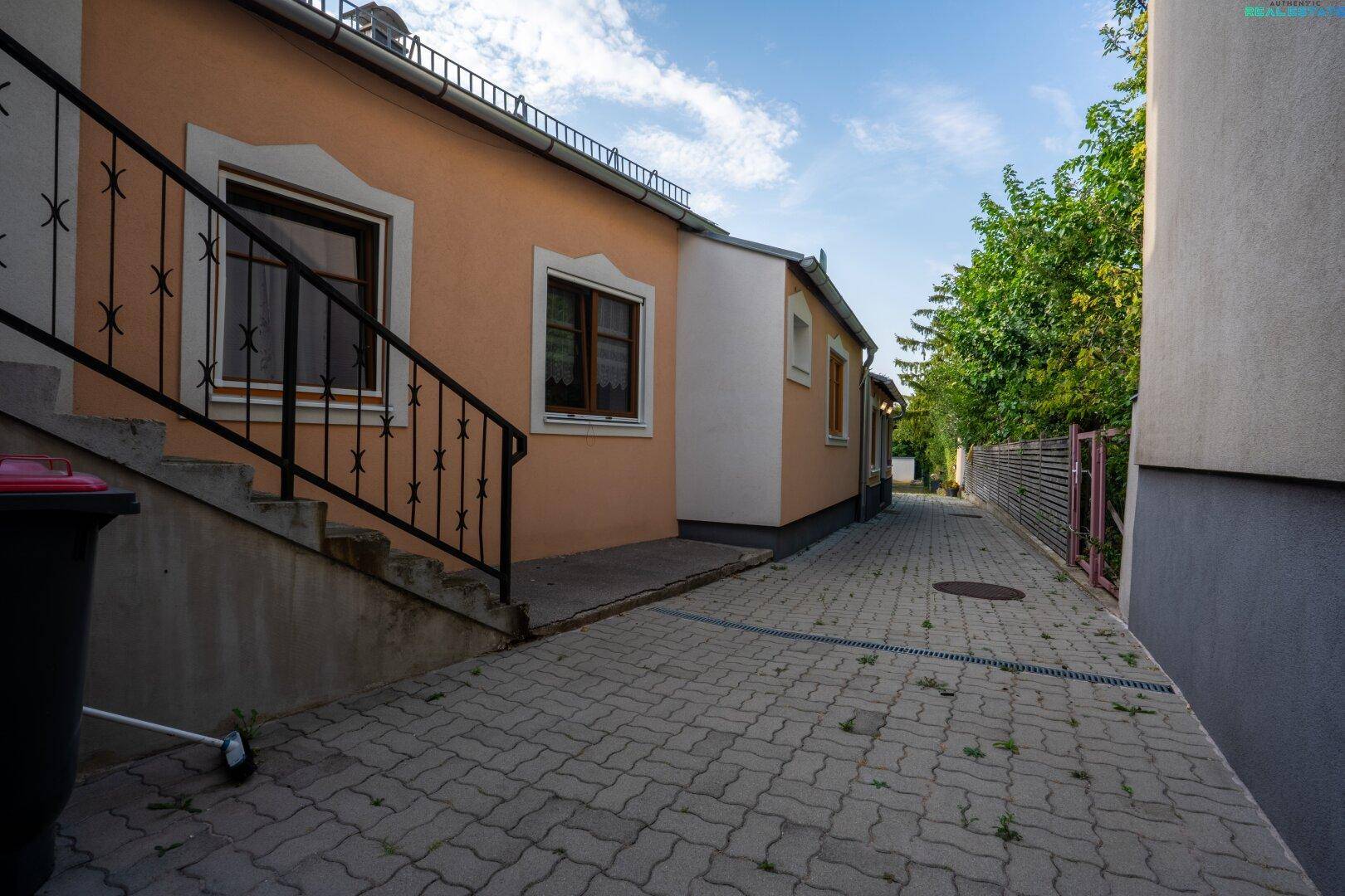 Gepflasterter Weg entlang der Hausfassade mit Außentreppe und Mülltonnen.