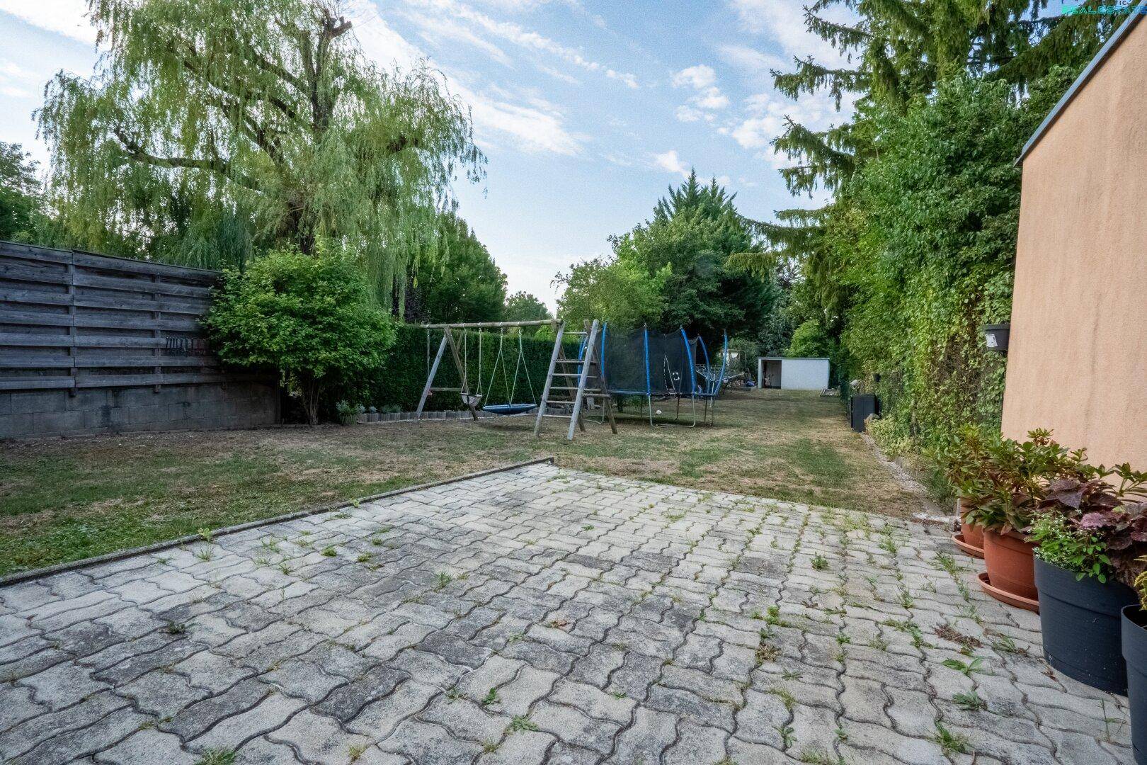 Gepflasterte Terrasse führt zu einem großen Garten mit Spielgeräten und Rasen.