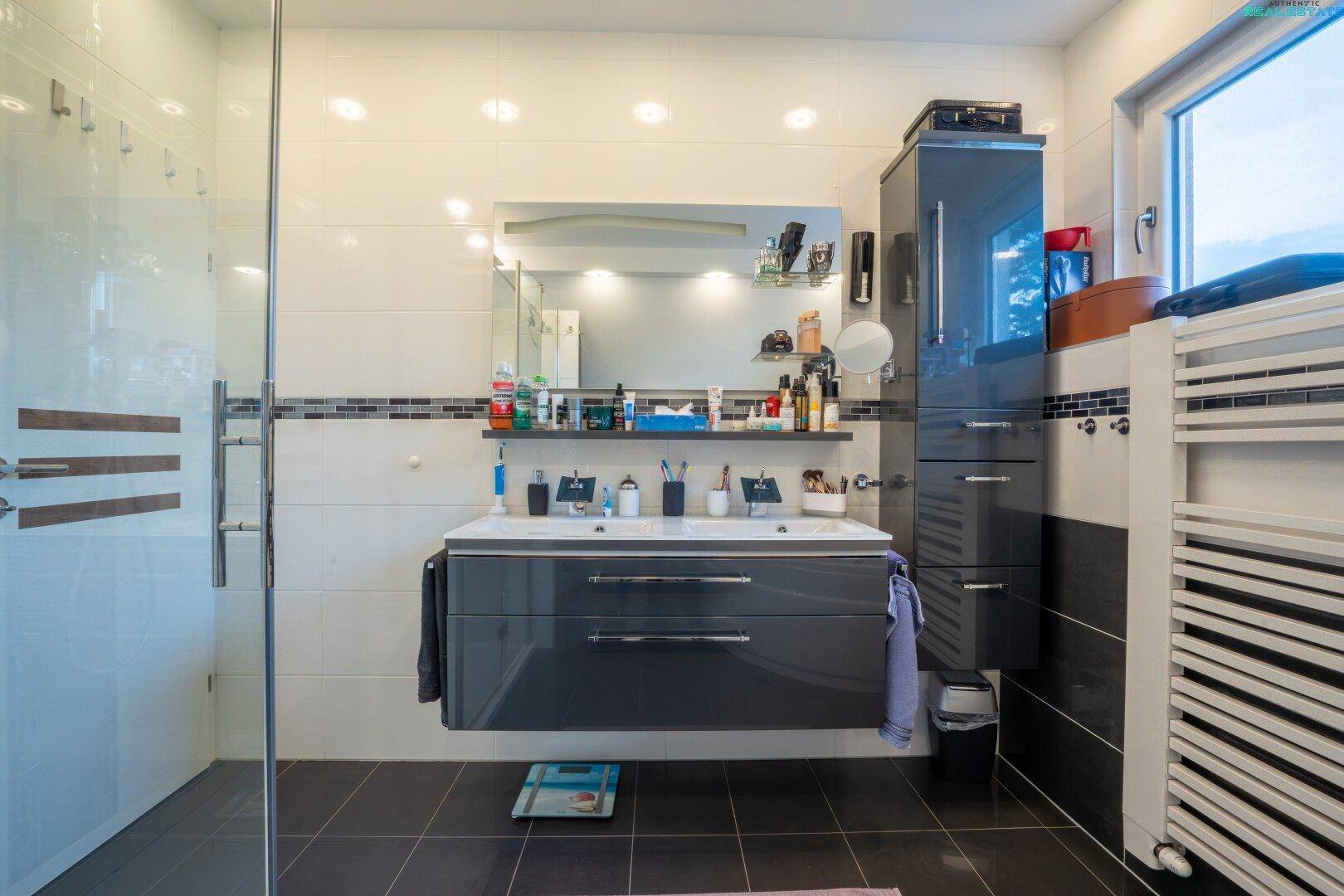 Modernes Badezimmer mit Doppelwaschbecken, Duschkabine und Fenster, in dunklen und hellen Fliesen.