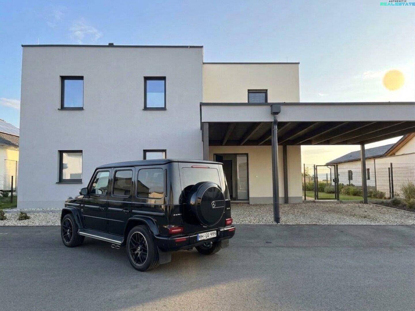 Moderne Hausfassade mit klaren Linien und einem Carport, davor ein geparktes Fahrzeug.
