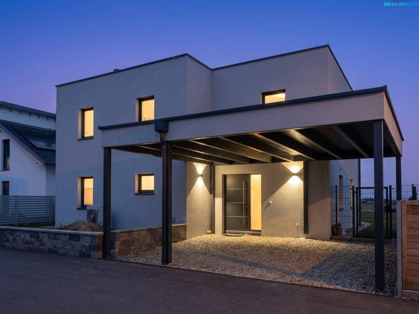Modernes Einfamilienhaus mit Carport bei Dämmerung, beleuchteter Eingangsbereich.
