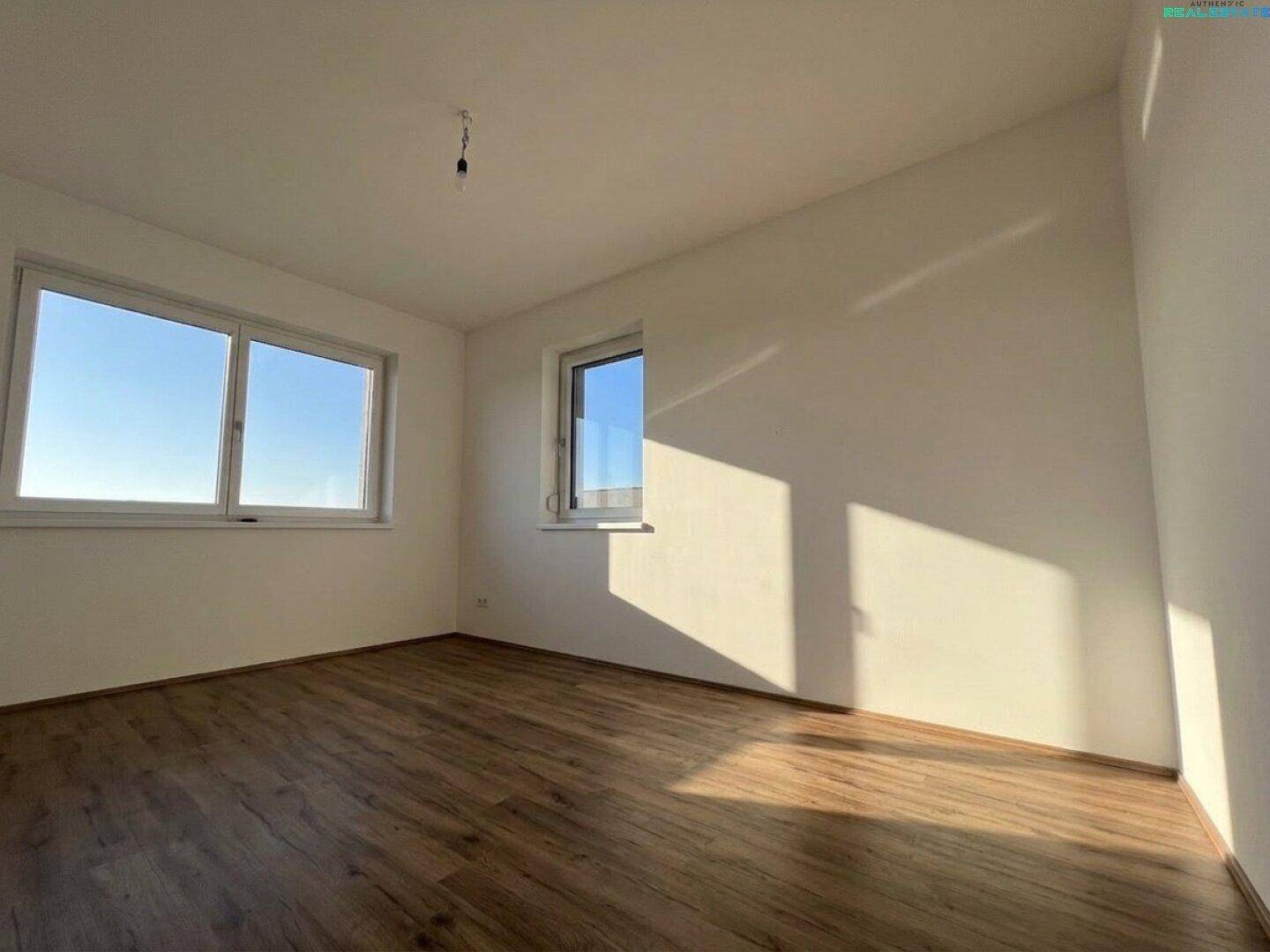 Helles, unmöbliertes Zimmer mit Laminatboden und großen Fenstern, sonnendurchflutet.