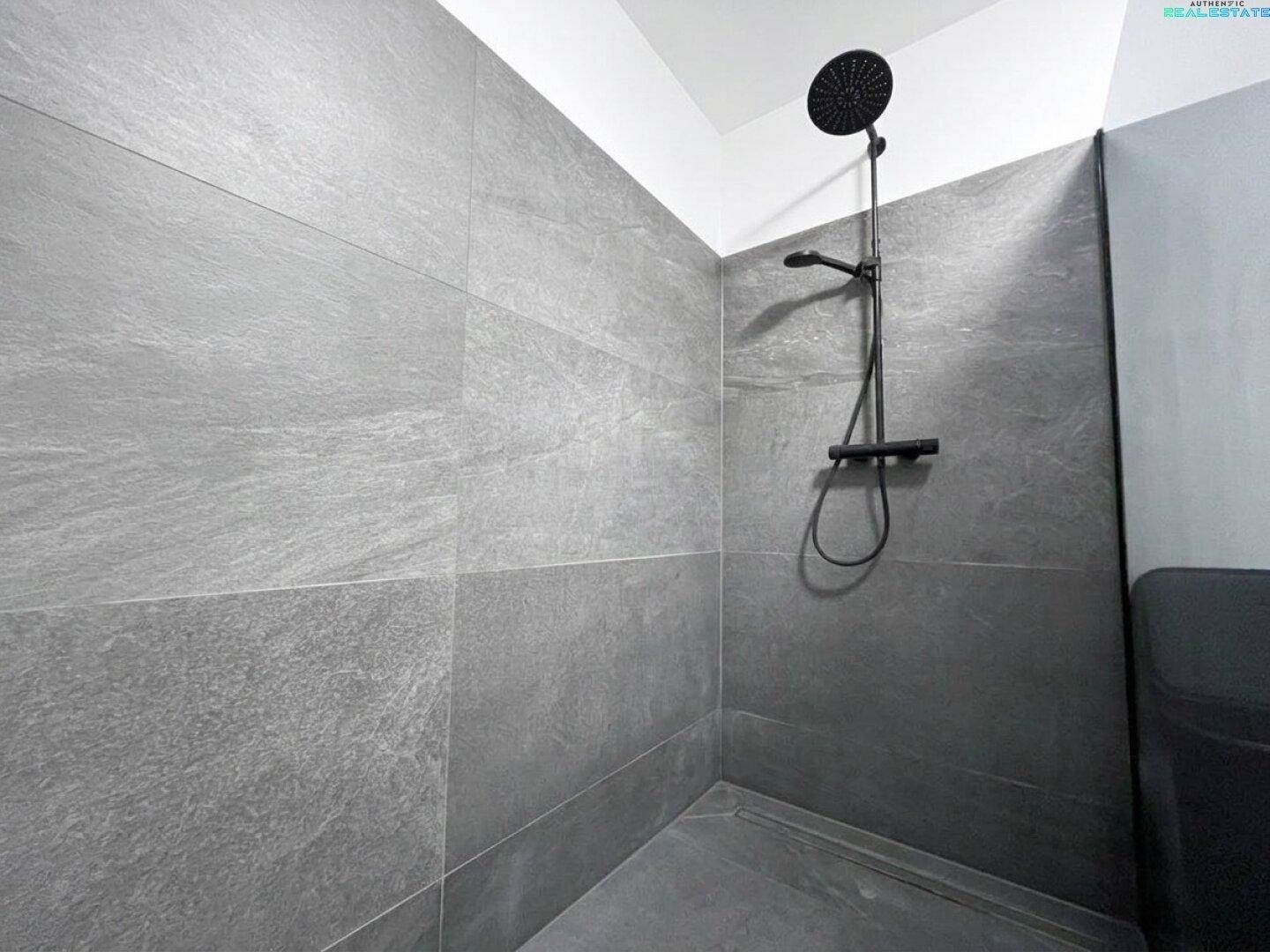 Duschbereich mit großformatigen grauen Fliesen und moderner schwarzer Regendusche.