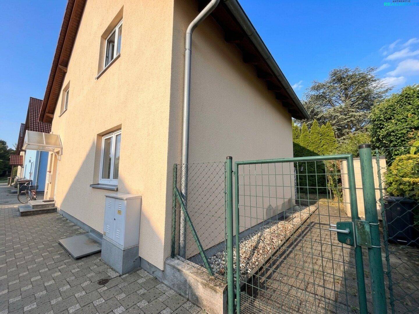 Seitenansicht des Hauses mit gepflastertem Weg und grünem Zaun unter blauem Himmel.