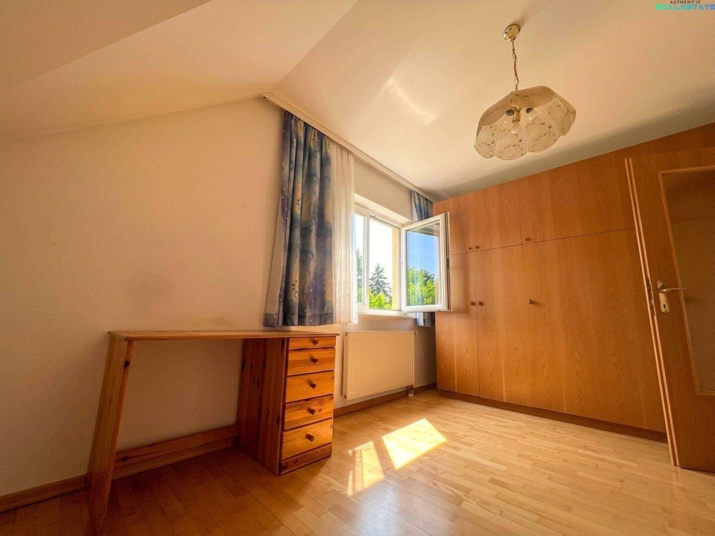Schlafzimmer mit Parkettboden, Holzschrank und Schreibtisch unter einem großen Fenster.