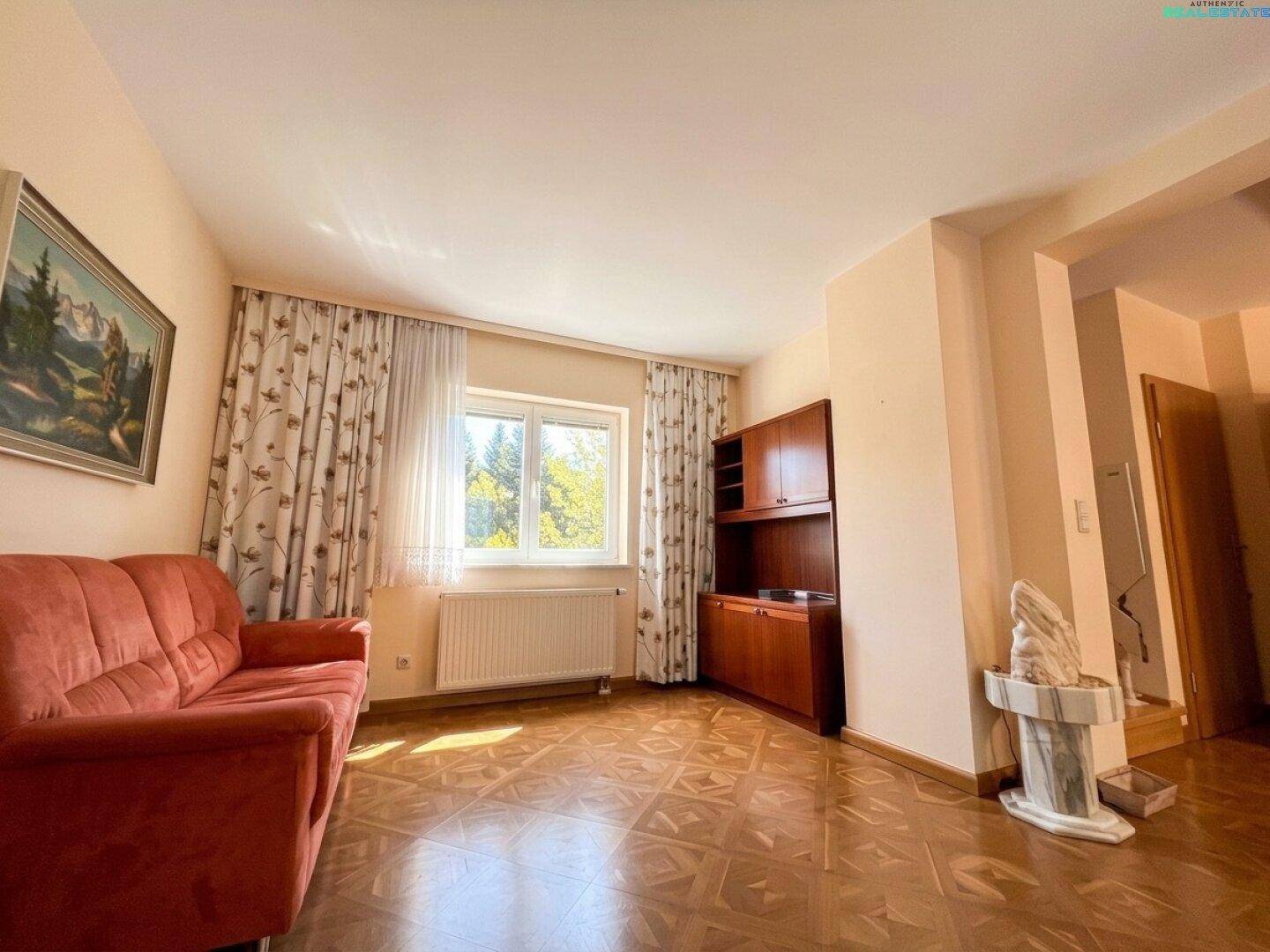 Geräumiges Zimmer mit Parkettboden, rotem Sofa und Fenster mit Blick ins Grüne.