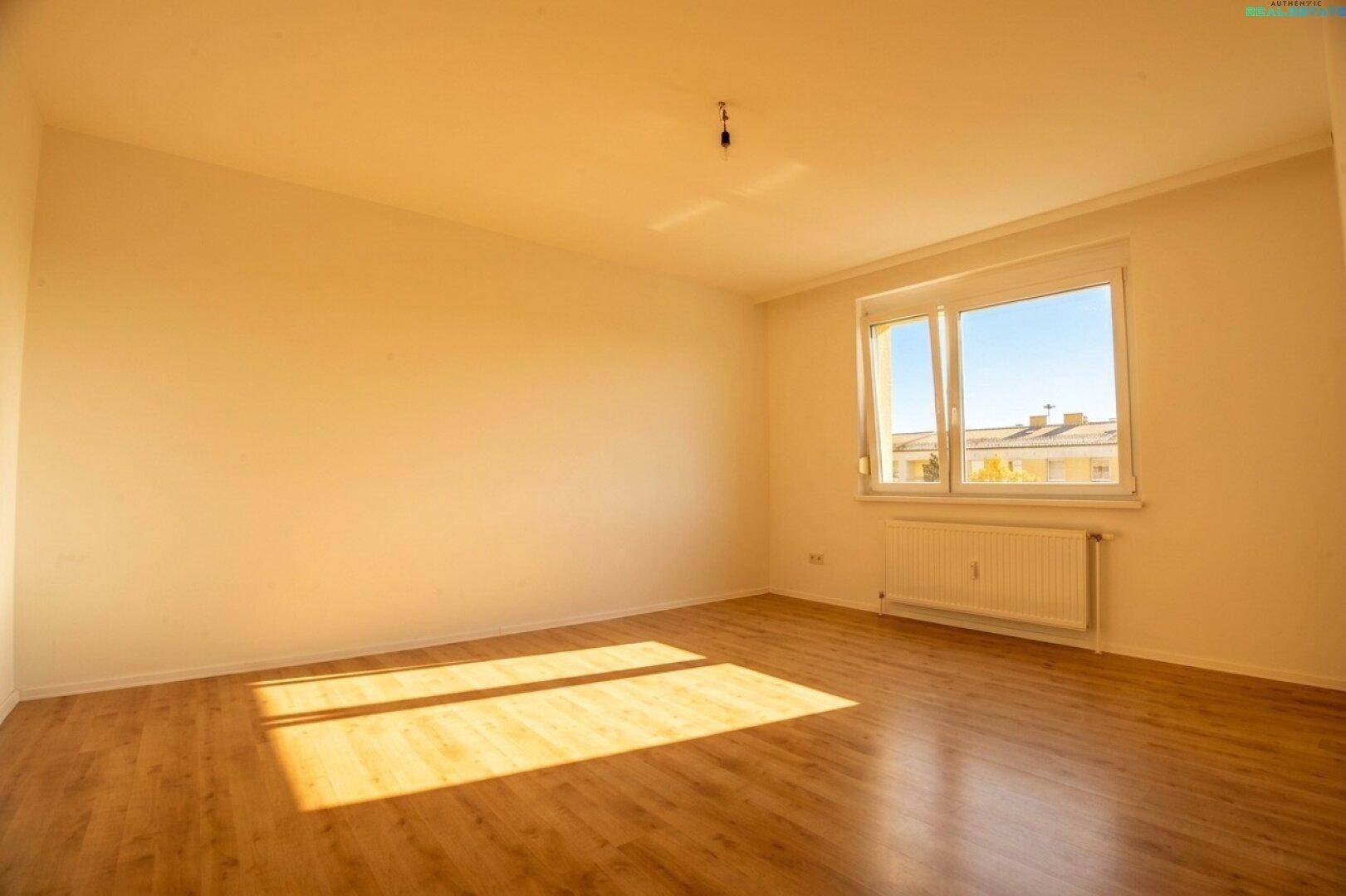 Leerer, heller Raum mit großem Fenster und Laminatboden, von Sonnenlicht durchflutet.