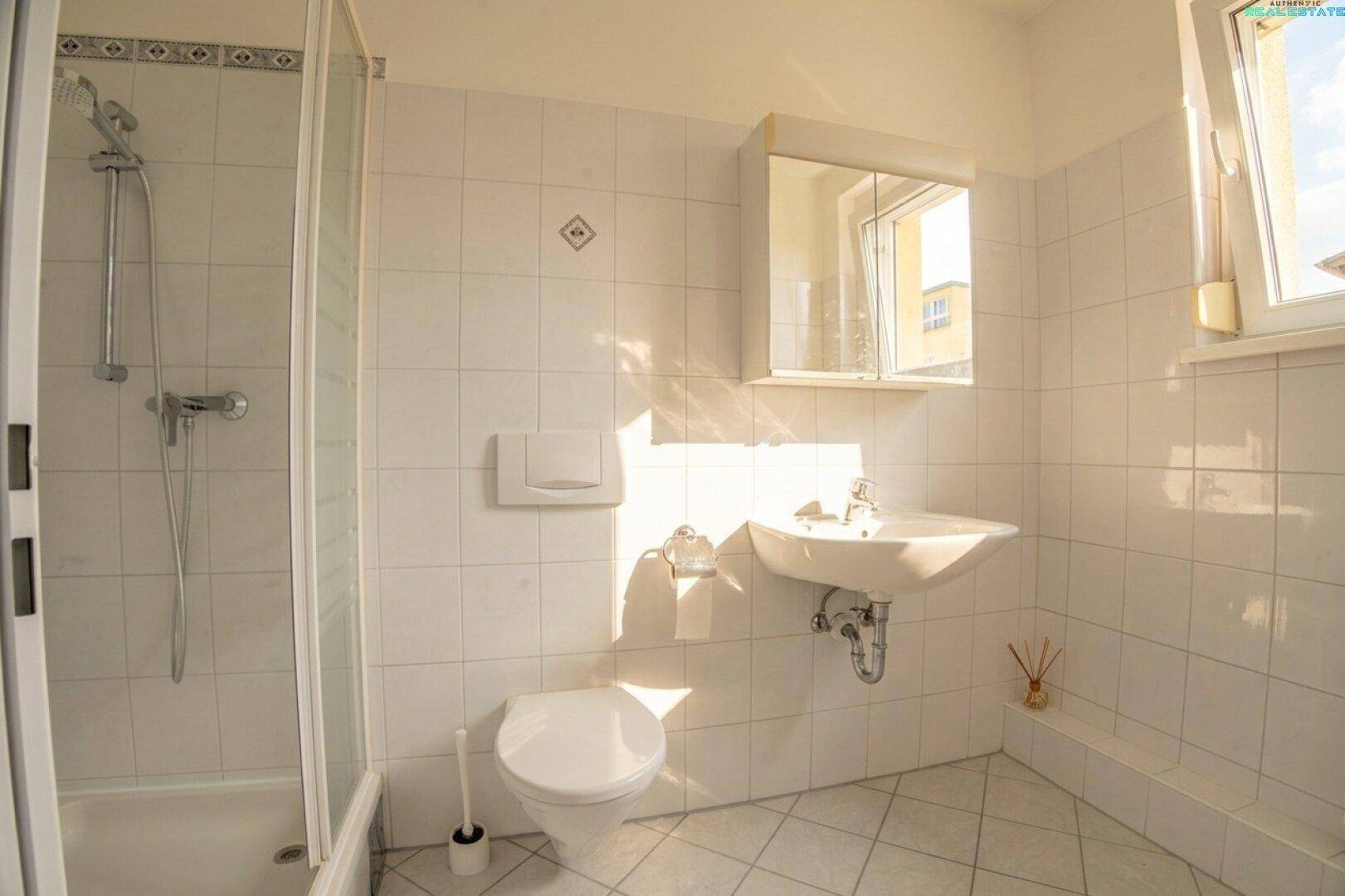 Modernes Badezimmer mit weißen Fliesen, Duschkabine, WC, Waschbecken und Spiegelschrank, hell beleuchtet.