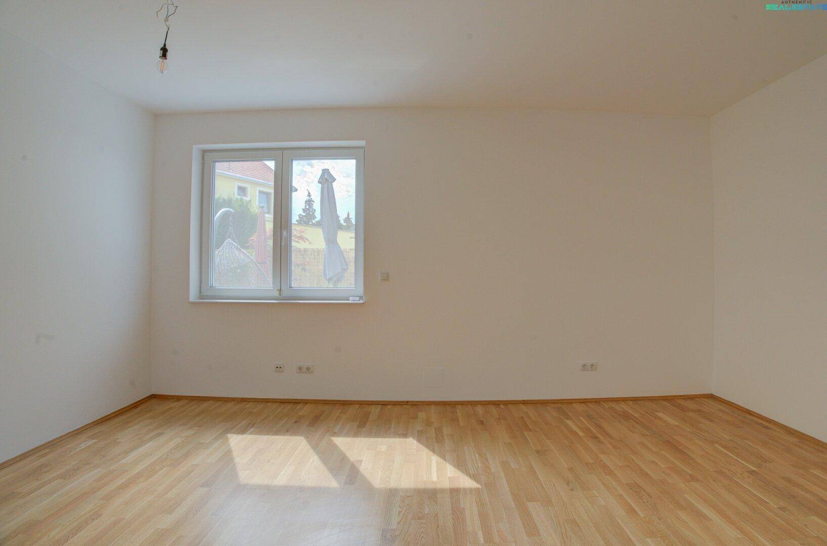 Leeres Zimmer mit hellem Parkettboden und einem Fenster, das Tageslicht hereinlässt.