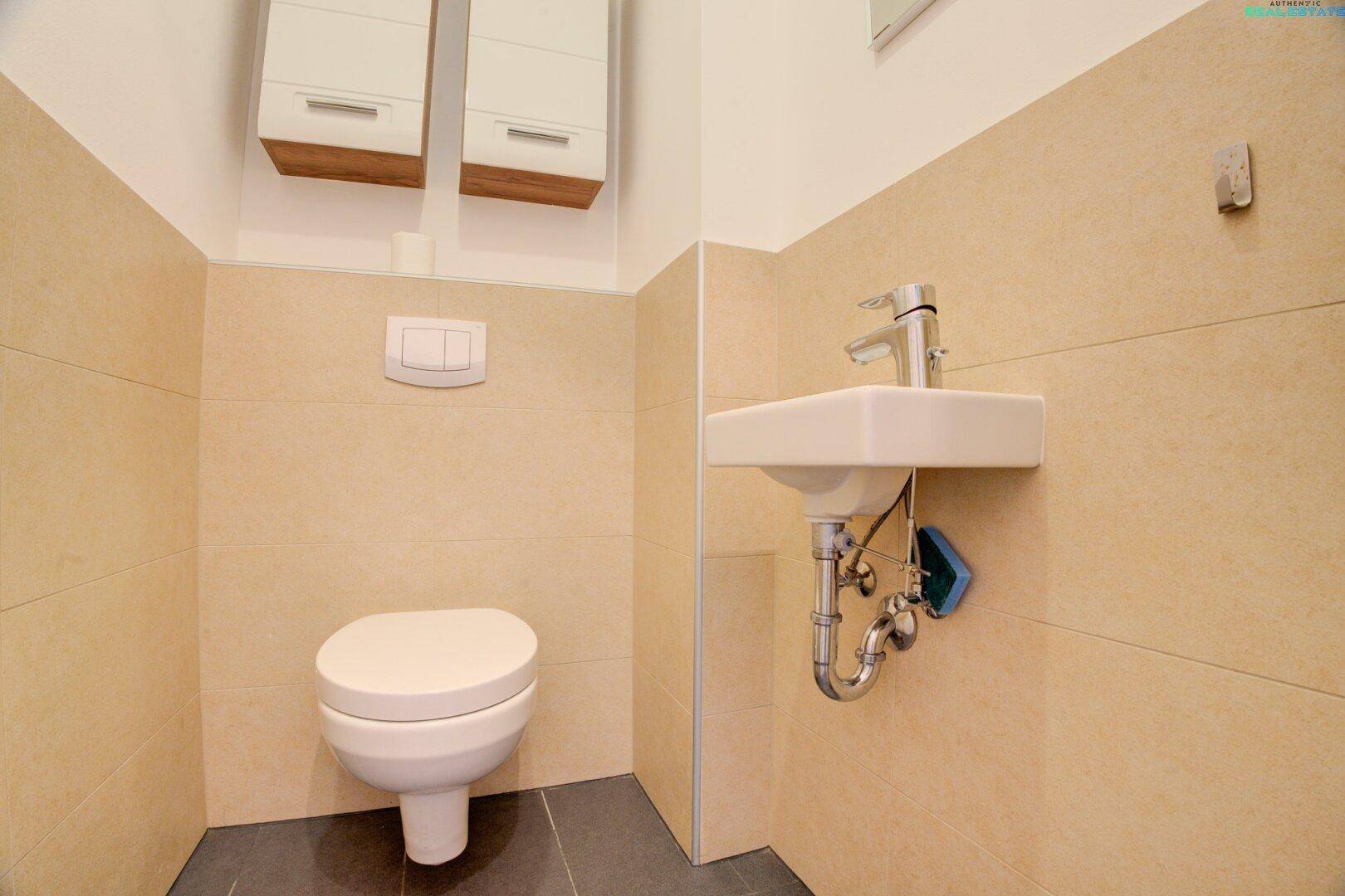 Gäste-WC mit wandhängender Toilette, kleinem Waschbecken und beigefarbenen Fliesen.