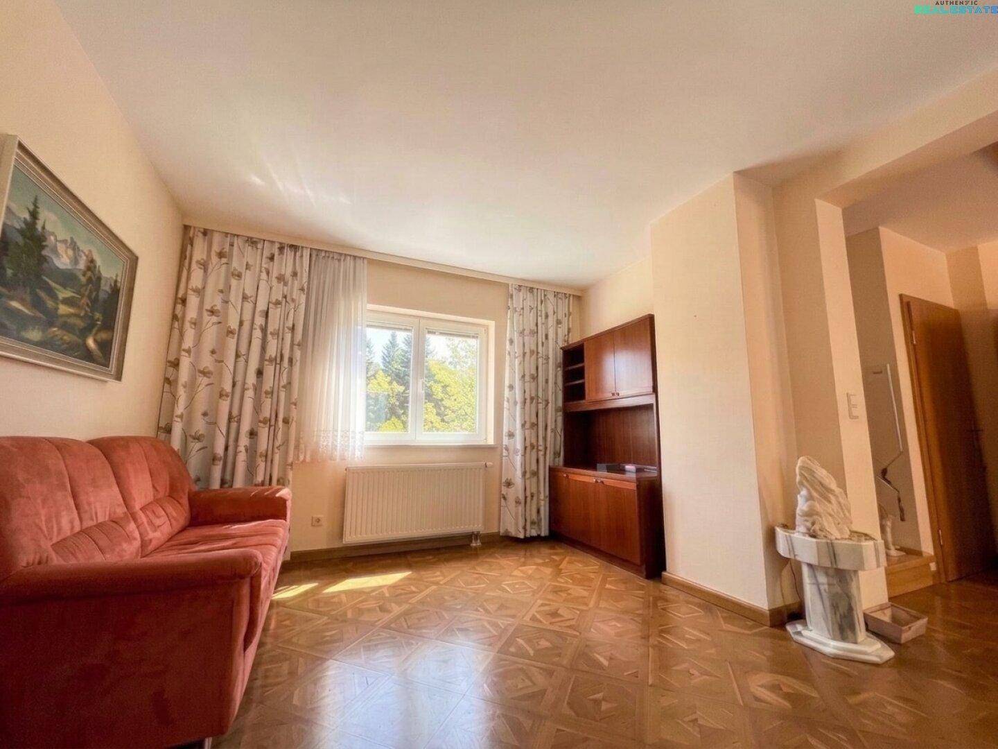 Helles Wohnzimmer mit Parkettboden, Sofa und Blick ins Grüne durch das Fenster.