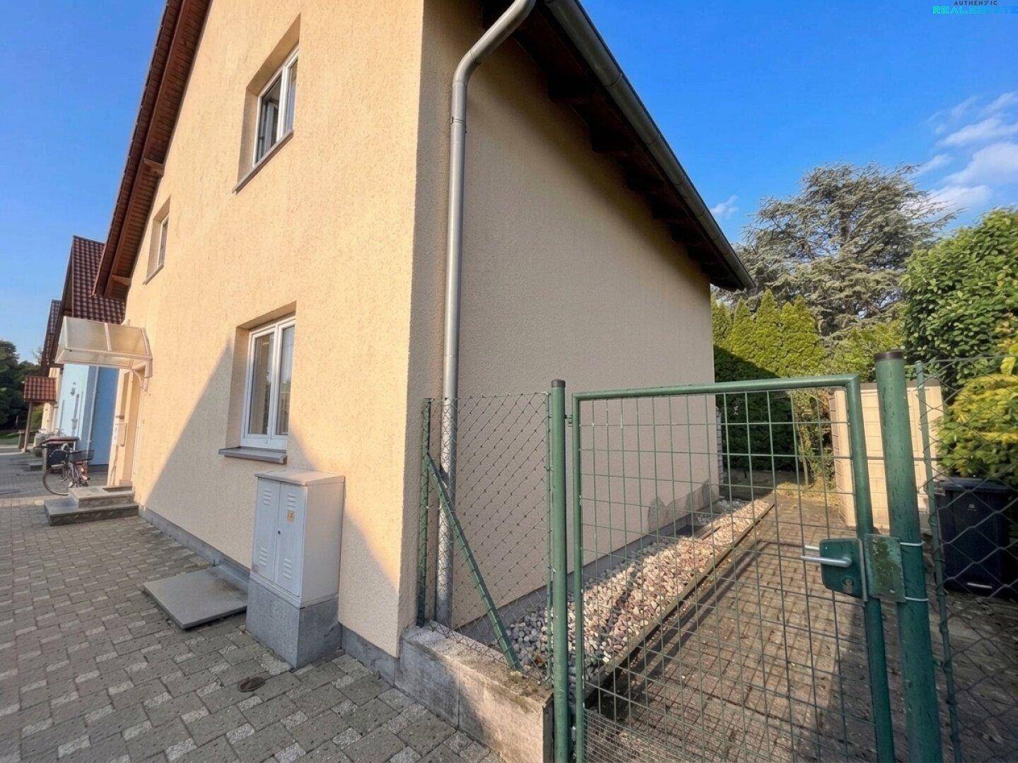 Seitenansicht des Hauses mit gepflastertem Weg, Zaun und Eingangsbereich im Sonnenlicht.