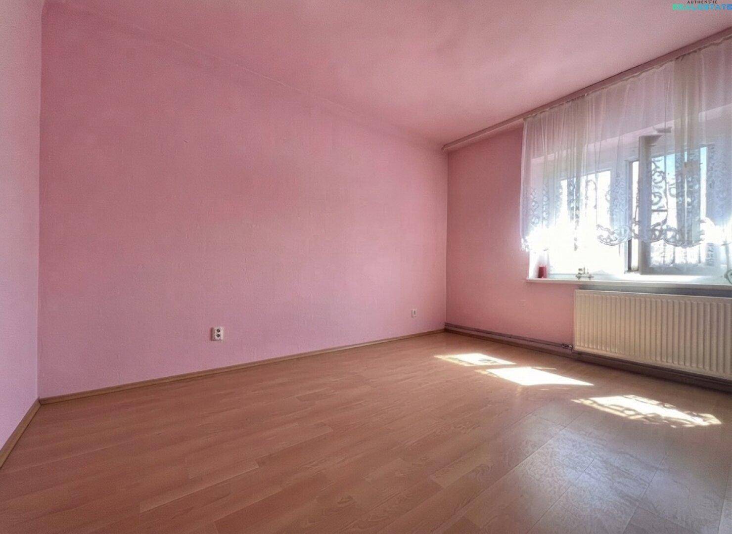 Leeres Zimmer mit rosa Wänden, hellem Laminatboden und einem Fenster mit Heizkörper.