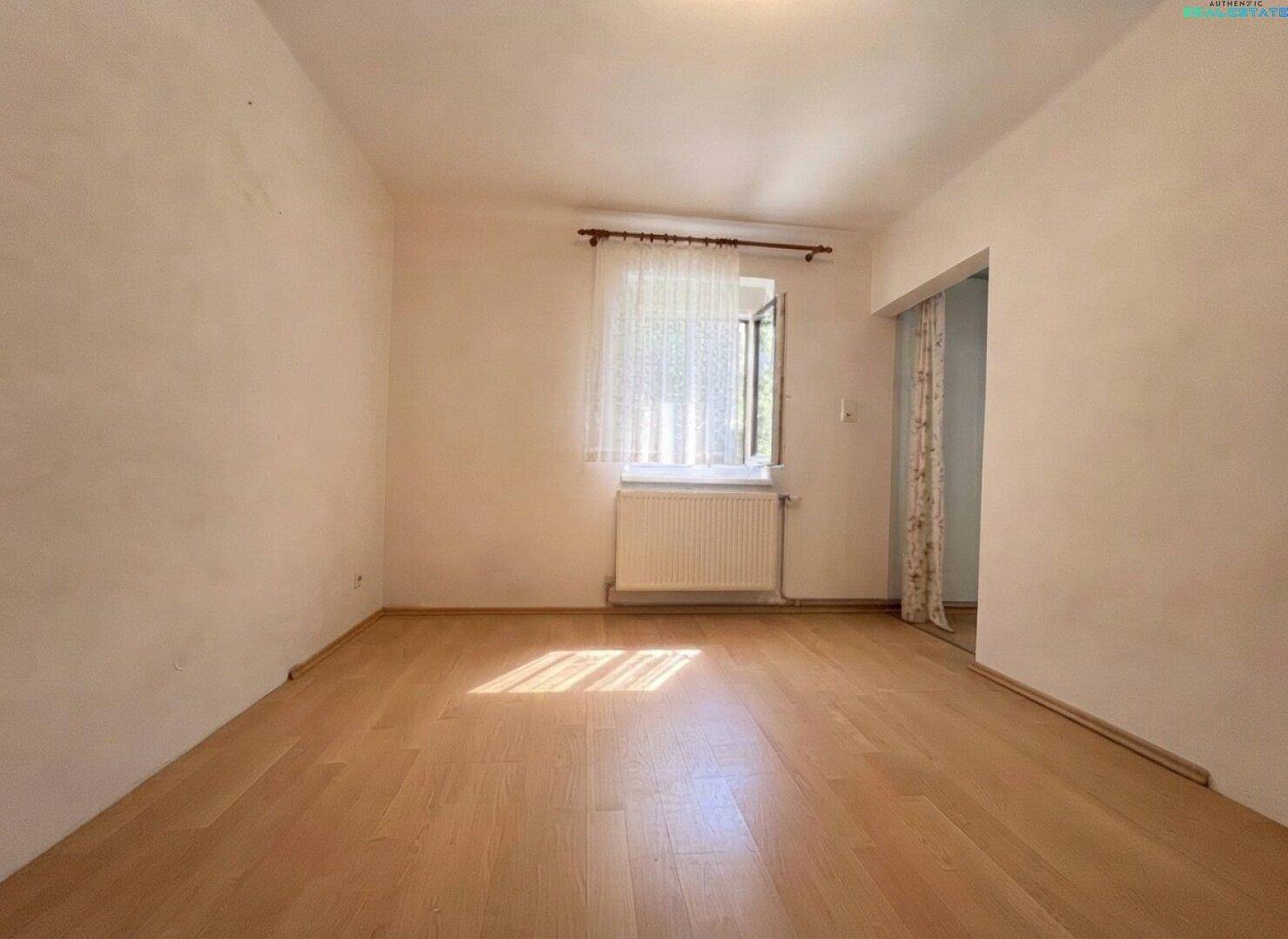 Schlichter Raum mit weißen Wänden, hellem Laminatboden und einem Fenster mit Blick ins Grüne.