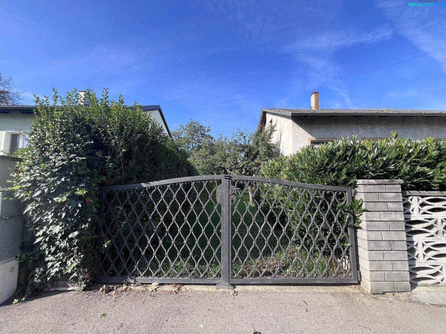 Geschmiedetes Eingangstor mit Hecken und Blick auf das Haus unter blauem Himmel.