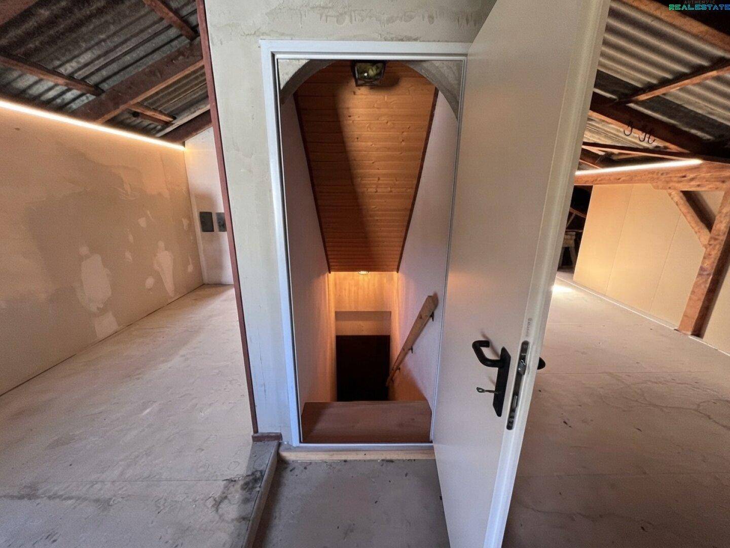 Unausgebauter Dachboden mit sichtbaren Holzbalken und Blick auf die Treppe.