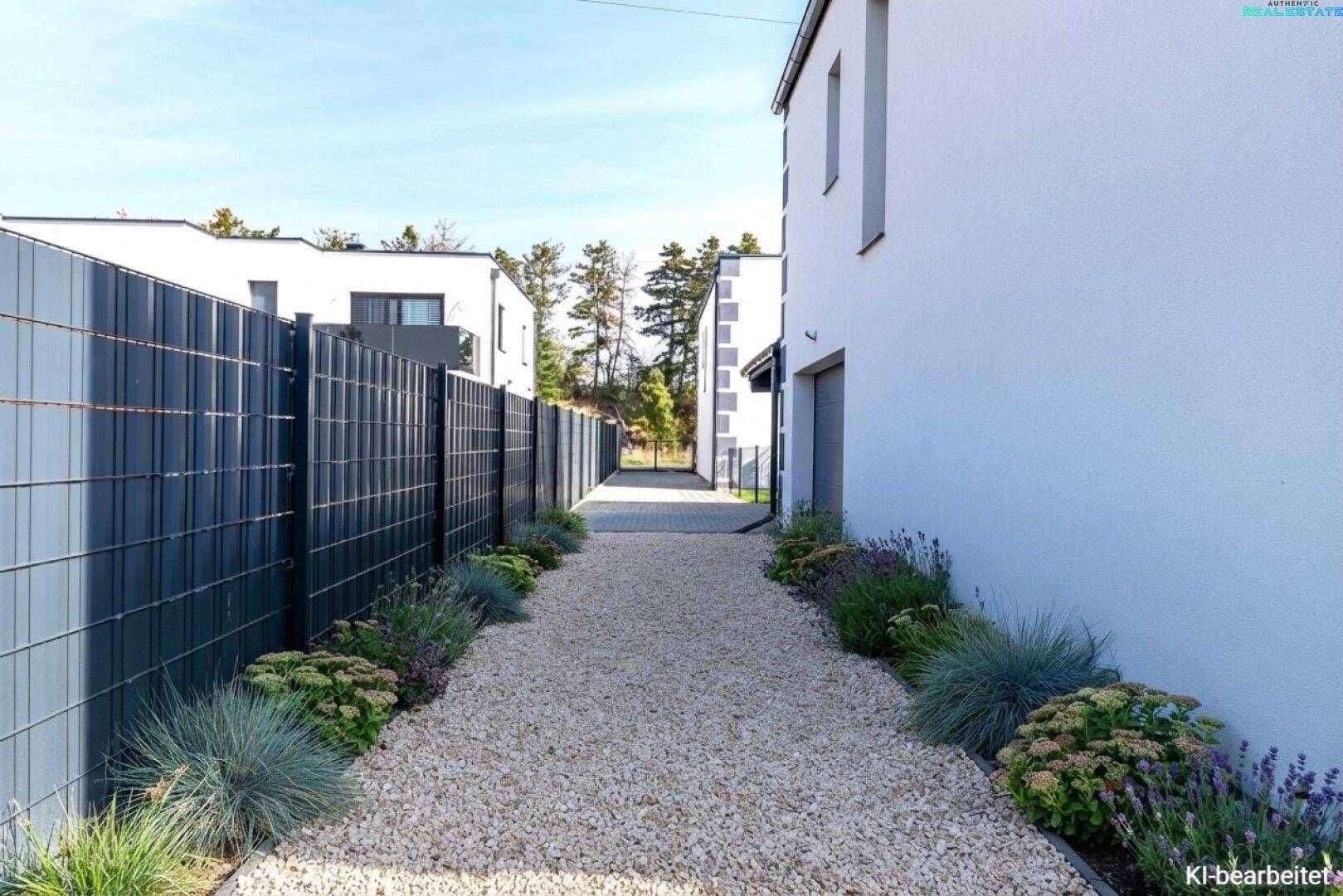 Kiesweg zwischen modernen Häusern mit gepflegten Beeten und Zaun.