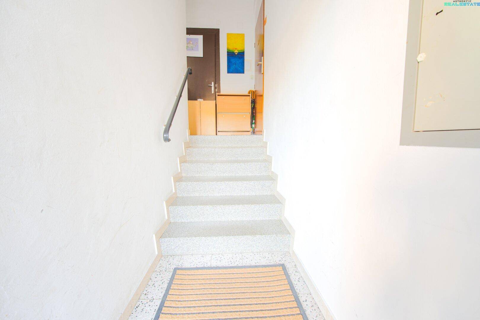 Helles Treppenhaus mit gefliestem Boden und Handlauf, das zu den oberen Etagen führt.