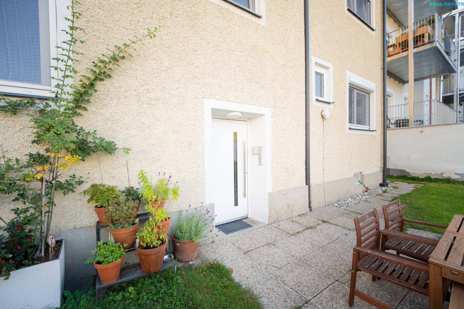 Eingangsbereich des Hauses mit weißer Tür, gepflasterter Terrasse und Topfpflanzen, einladend gestaltet.
