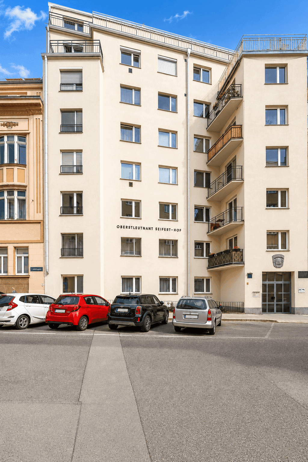 Mehrstöckiges Wohngebäude mit Balkonen und Parkplätzen davor unter blauem Himmel.