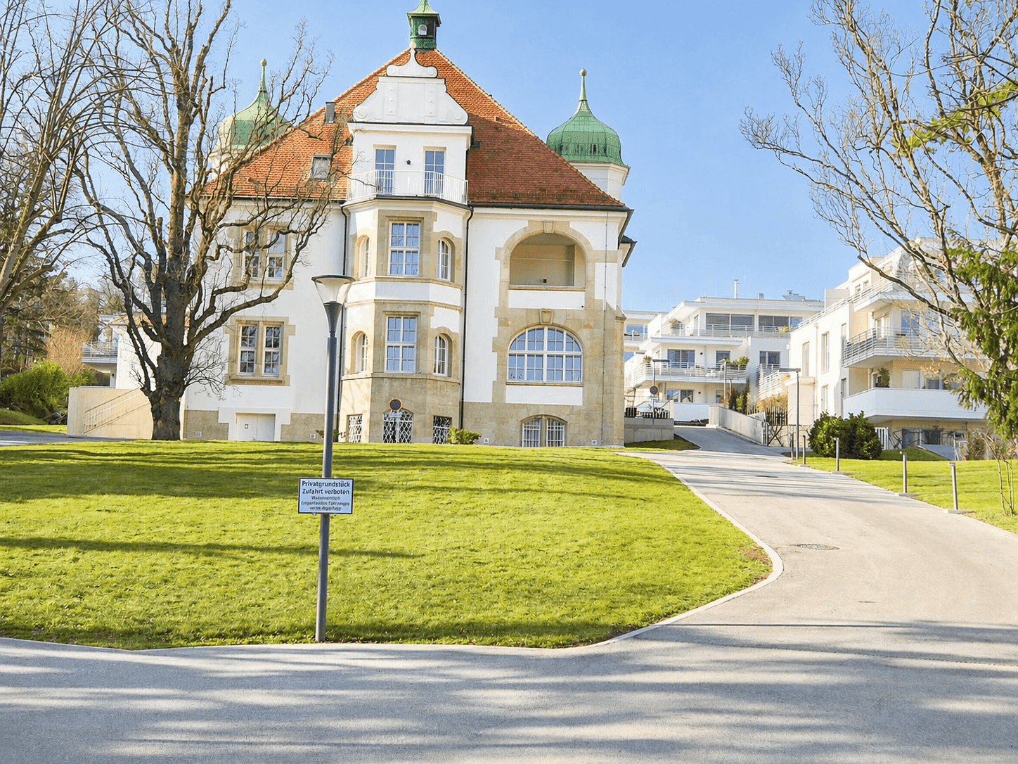 Historisches Gebäude mit markantem Dach und gepflegter Grünfläche, umgeben von moderner Bebauung.