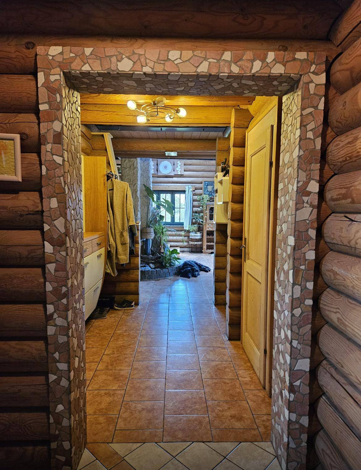 Blick durch einen Flur im Blockhaus mit Holzstammwänden, Fliesenboden und einem dekorativen Steinbogen.