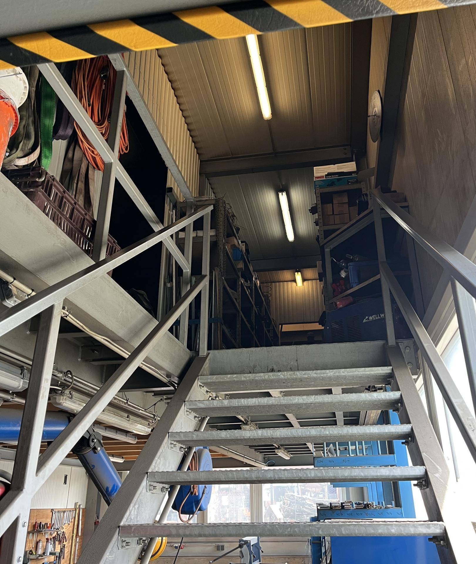 Metalltreppe führt zu einer oberen Lagerfläche mit Regalen und industrieller Beleuchtung.