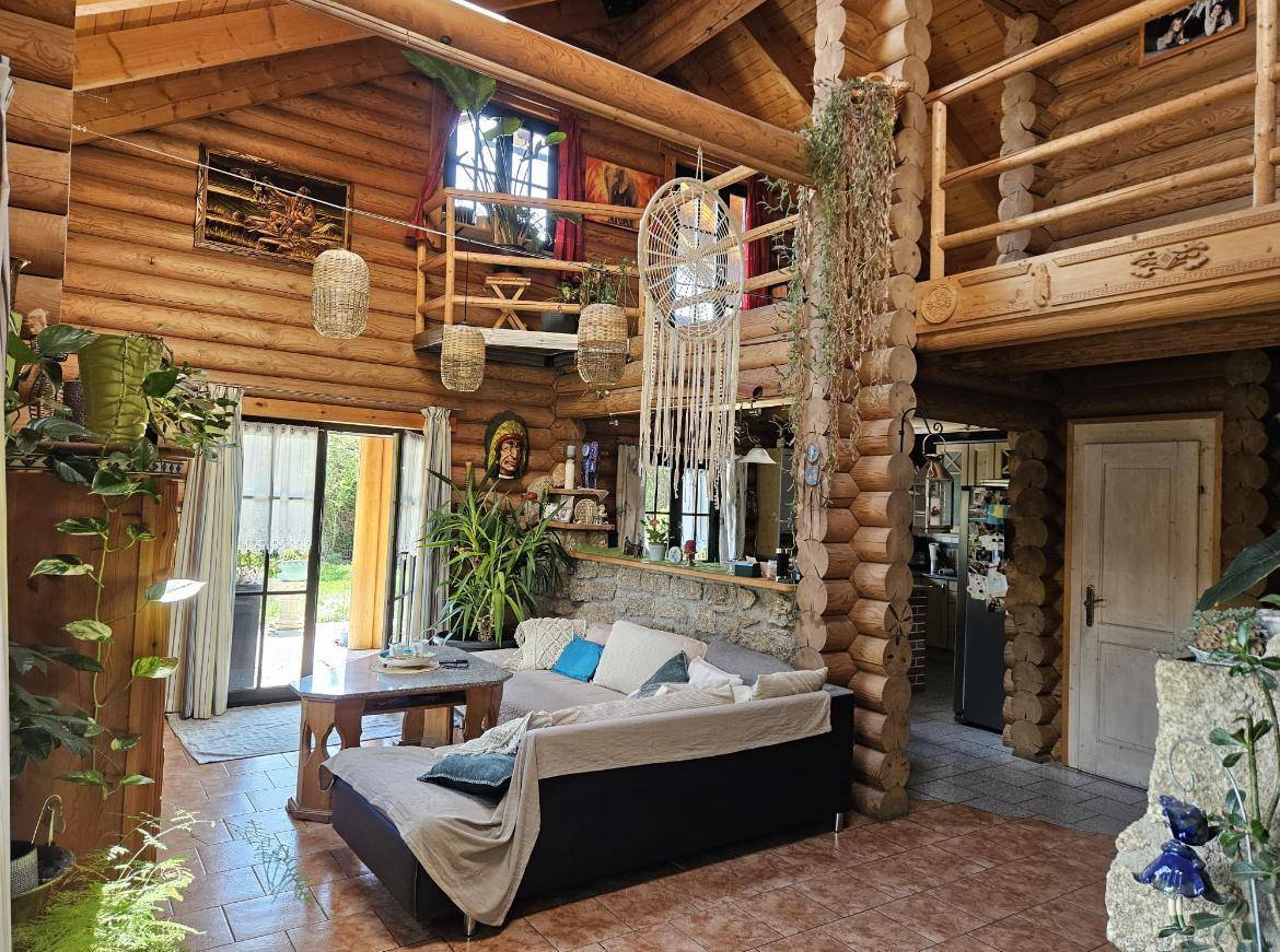 Wohnzimmer im Blockhaus mit hohen Decken, sichtbaren Holzstämmen, Fliesenboden und Blick auf eine Galerie.