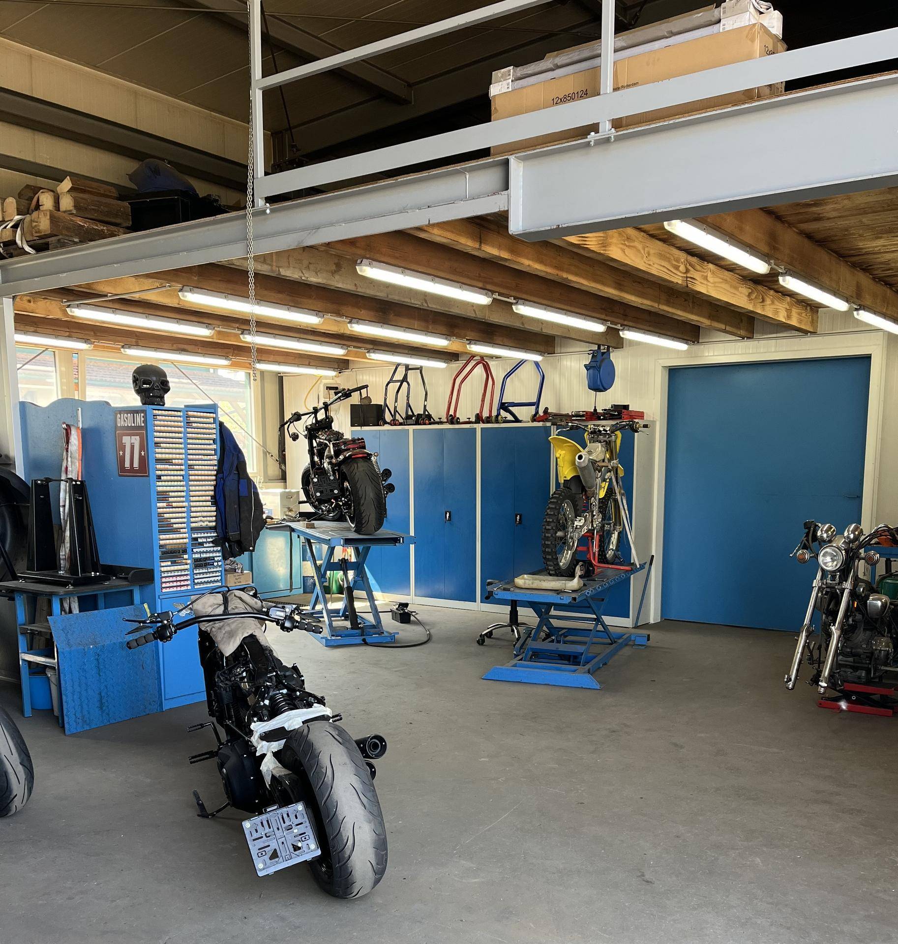 Werkstatt mit mehreren Motorrädern auf Hebebühnen, blauen Werkzeugschränken und sichtbaren Holzdeckenbalken.