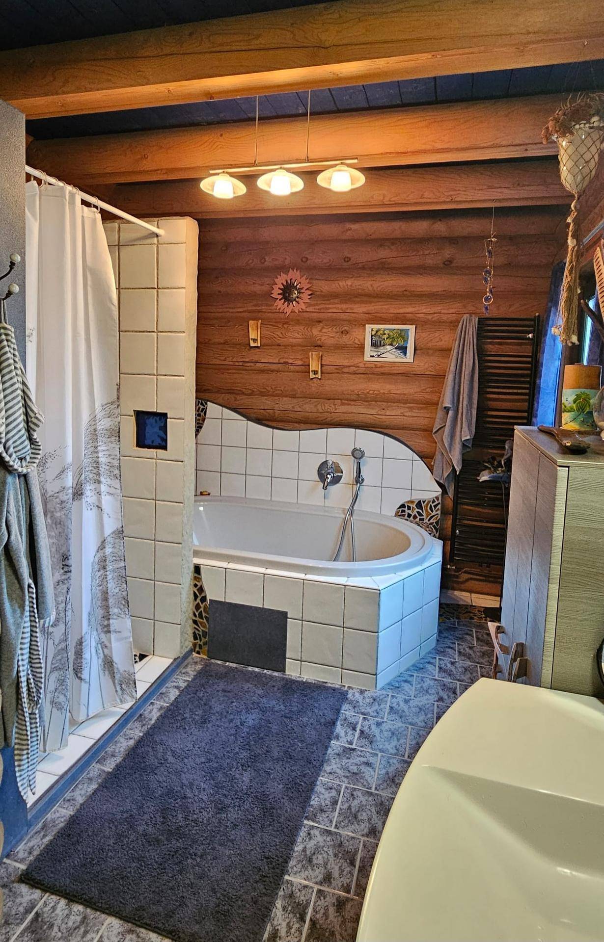 Badezimmer im Blockhaus mit Eckbadewanne, Duschbereich, gefliesten Wänden und Holzstammwänden.