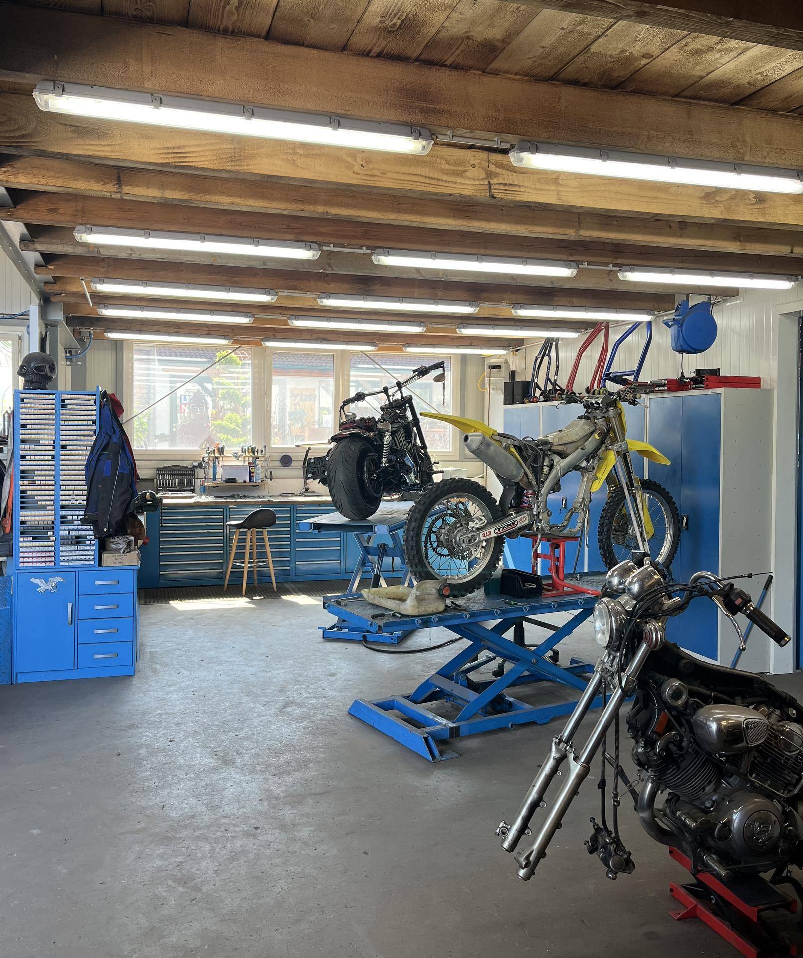 Werkstatt mit mehreren Motorrädern auf Hebebühnen, blauen Werkzeugschränken und sichtbaren Holzdeckenbalken.