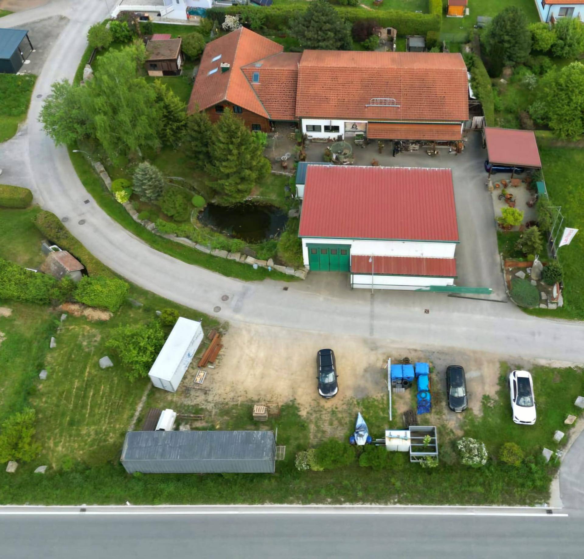 Luftaufnahme des Anwesens mit Haupthaus, großem Nebengebäude, Teich und einem Parkplatz mit mehreren Fahrzeugen.