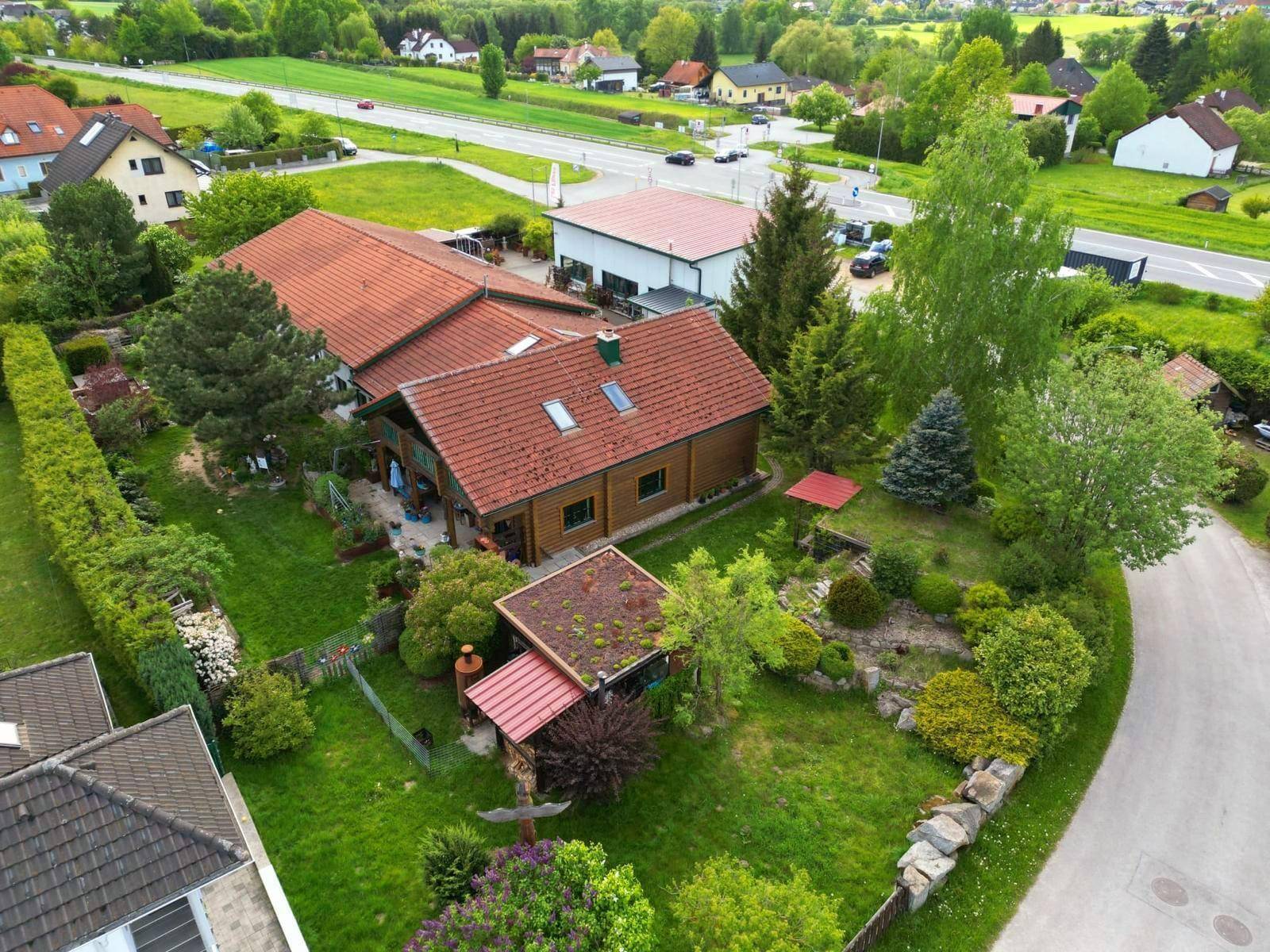 Luftaufnahme des Anwesens mit Blockhaus, Nebengebäude und der umliegenden ländlichen Landschaft mit Straße und weiteren Häusern.