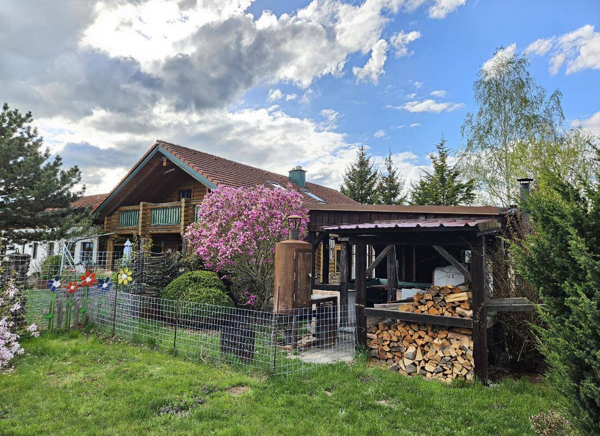 Außenansicht des Blockhauses mit einem überdachten Holzschuppen, einem blühenden Rhododendron und einem Gartenzaun.