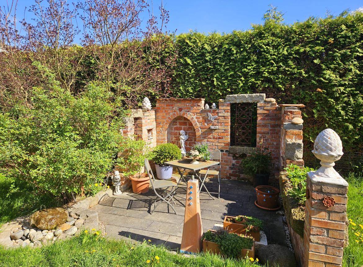 Gartenbereich mit kleiner gepflasterter Terrasse, einer Ziegelmauer und Gartenmöbeln unter blauem Himmel.