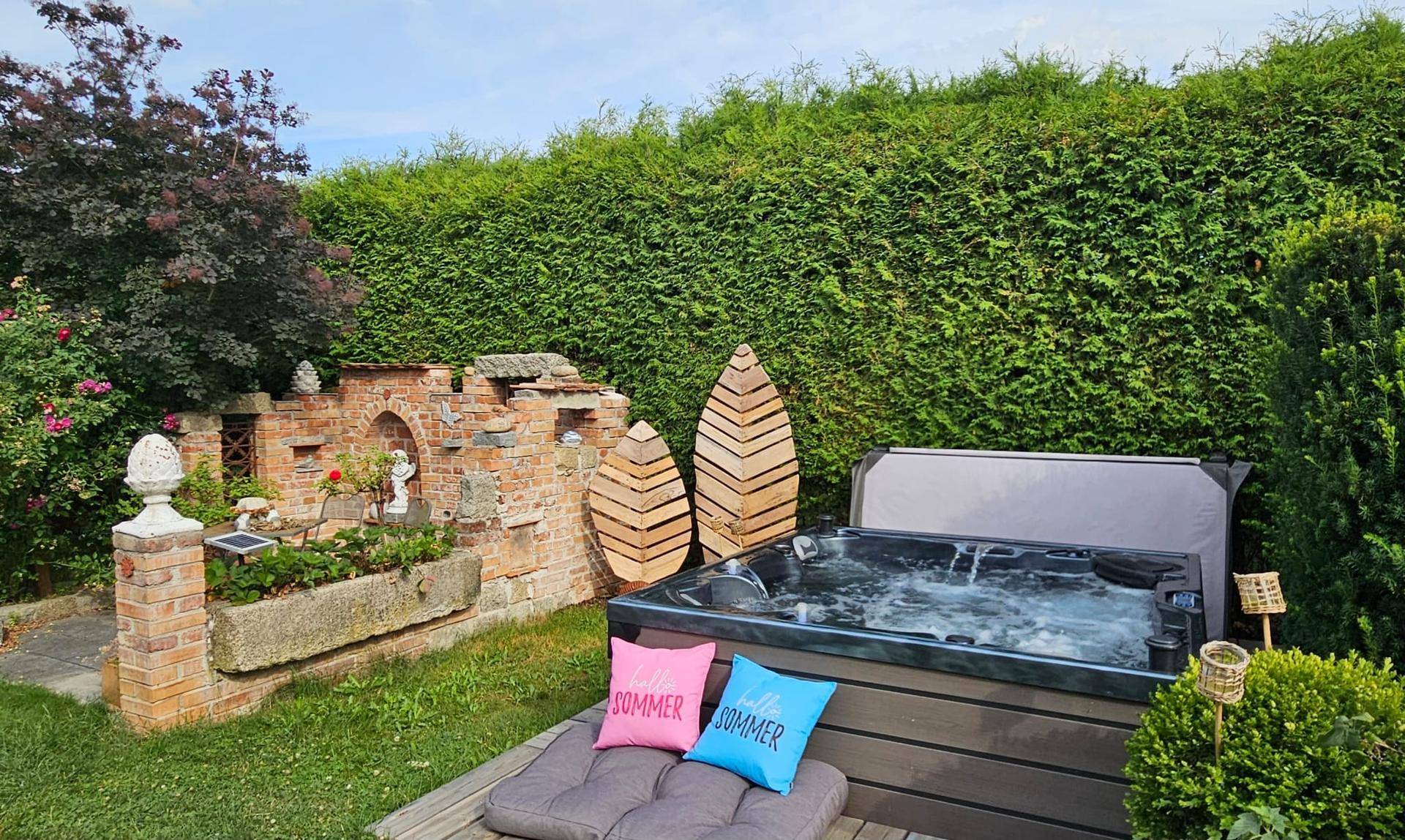 Gartenbereich mit einem sprudelnden Whirlpool, umgeben von einer hohen Hecke und dekorativen Elementen.