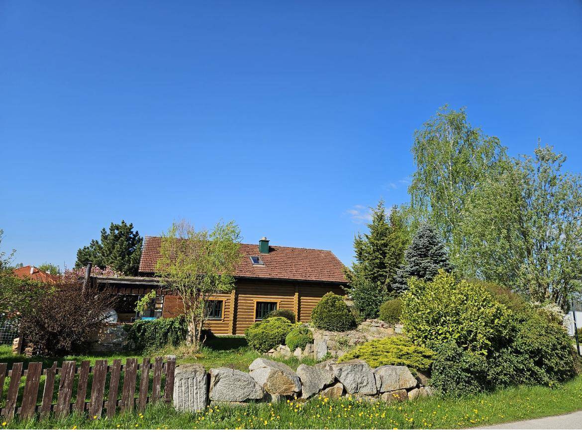 Blockhaus von Weitem hinter einem Holzzaun und einer Steinmauer, umgeben von Bäumen unter klarem blauem Himmel.