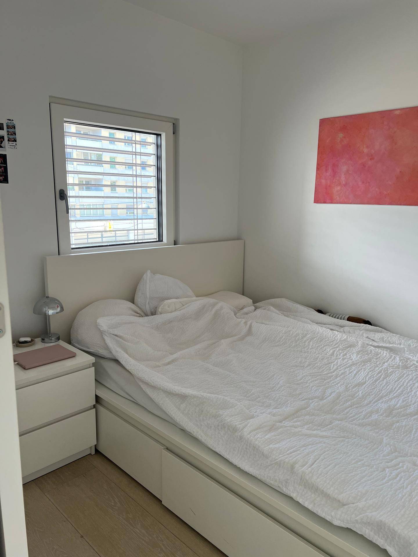 Helles Schlafzimmer mit weißem Bett, Nachttisch und einem Fenster mit Blick nach draußen.