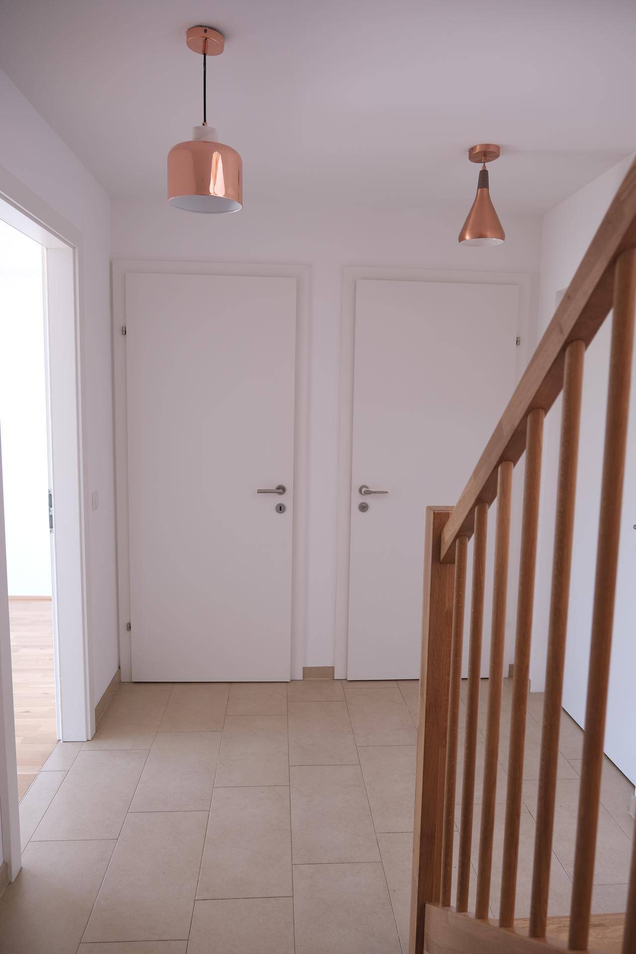 Schlichter Flur mit hellen Fliesen, zwei Türen und einem Holzgeländer der Treppe.