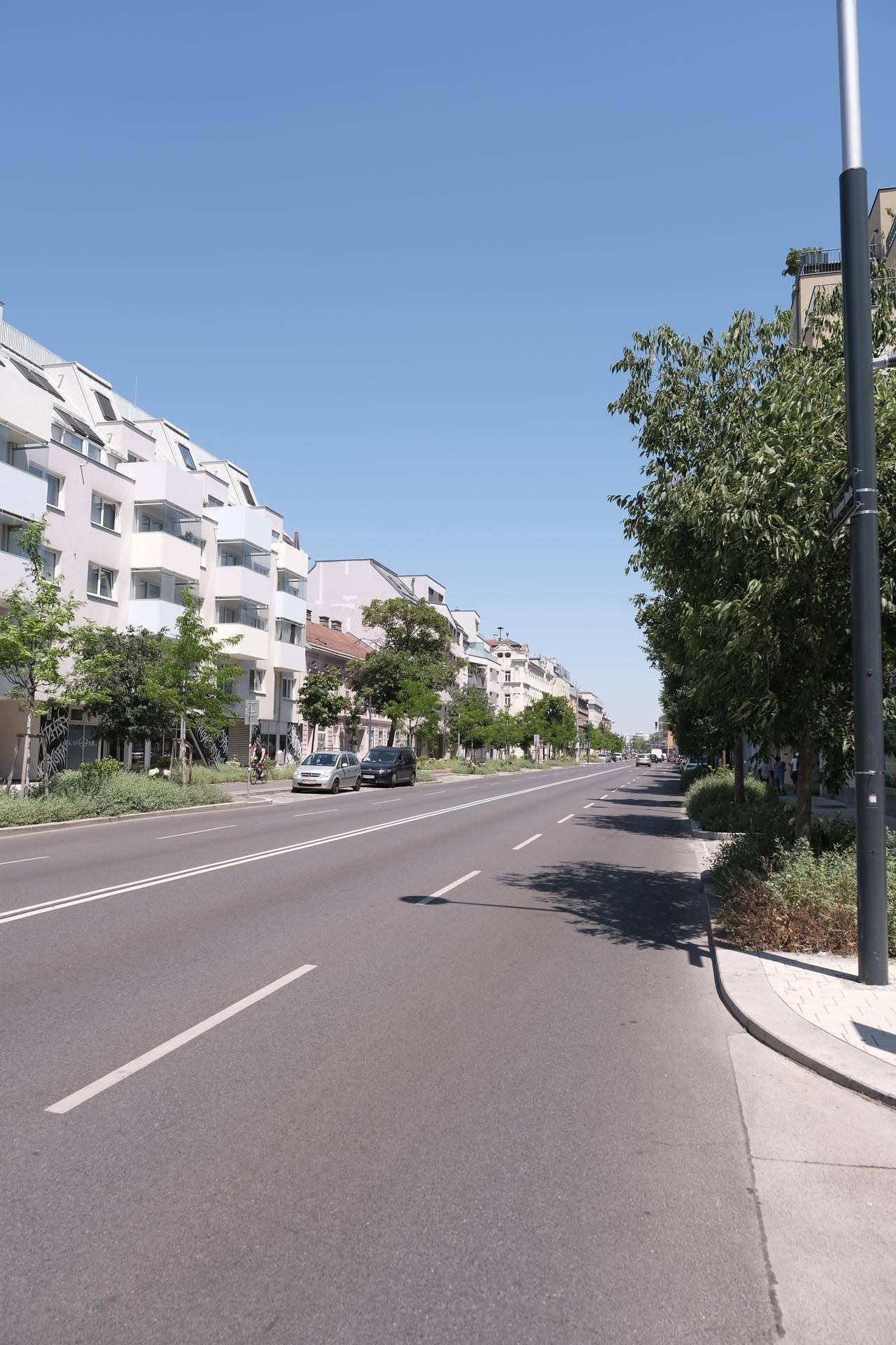 Breite Wohnstraße mit modernen Gebäuden, Bäumen und klarem blauem Himmel.