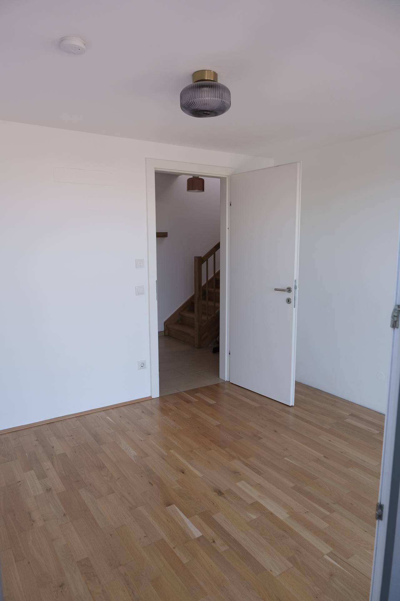 Helles, leeres Schlafzimmer mit Holzboden und Zugang zum Flur mit der Holztreppe.