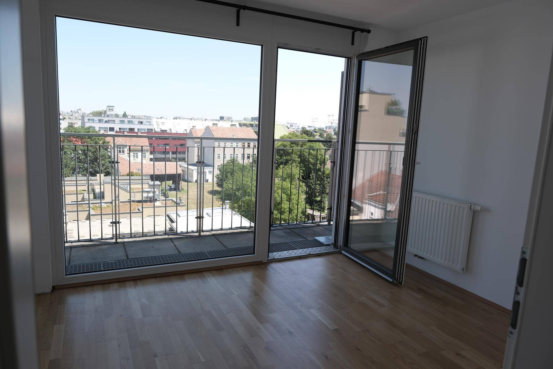 Heller Raum mit Holzboden, großem Fenster und Zugang zu einem Balkon mit Stadtblick.