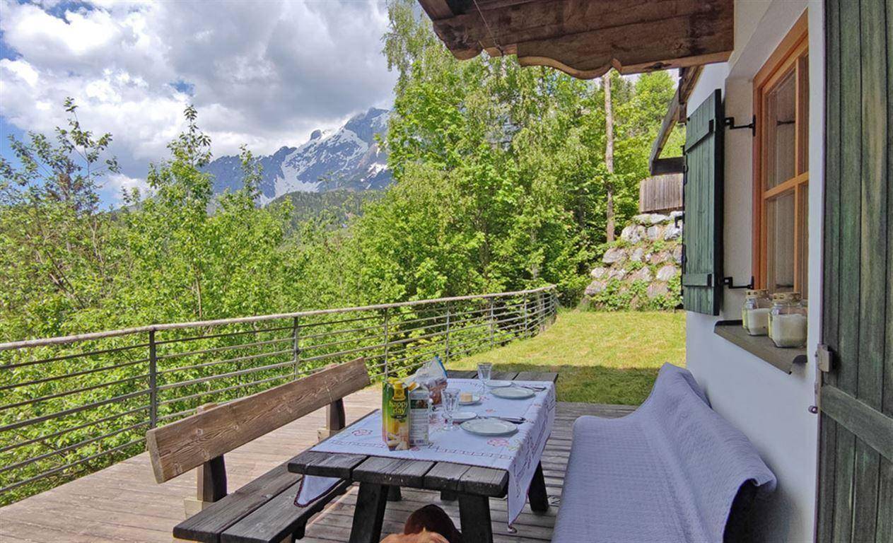 Gemütliche Holzterrasse mit gedecktem Tisch und Sitzbänken, umgeben von Grün und Bergblick.