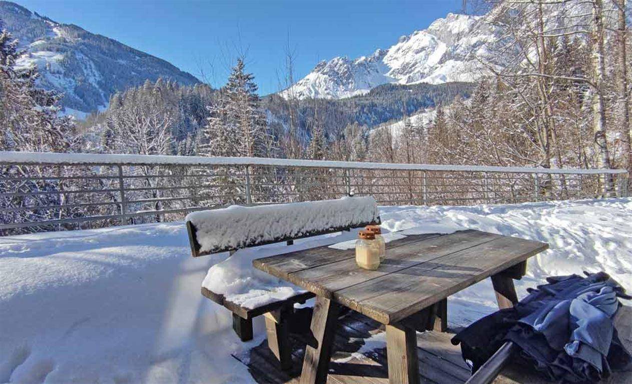 Rustikale Holzmöbel auf einer verschneiten Terrasse mit beeindruckendem Blick auf die winterliche Bergwelt.